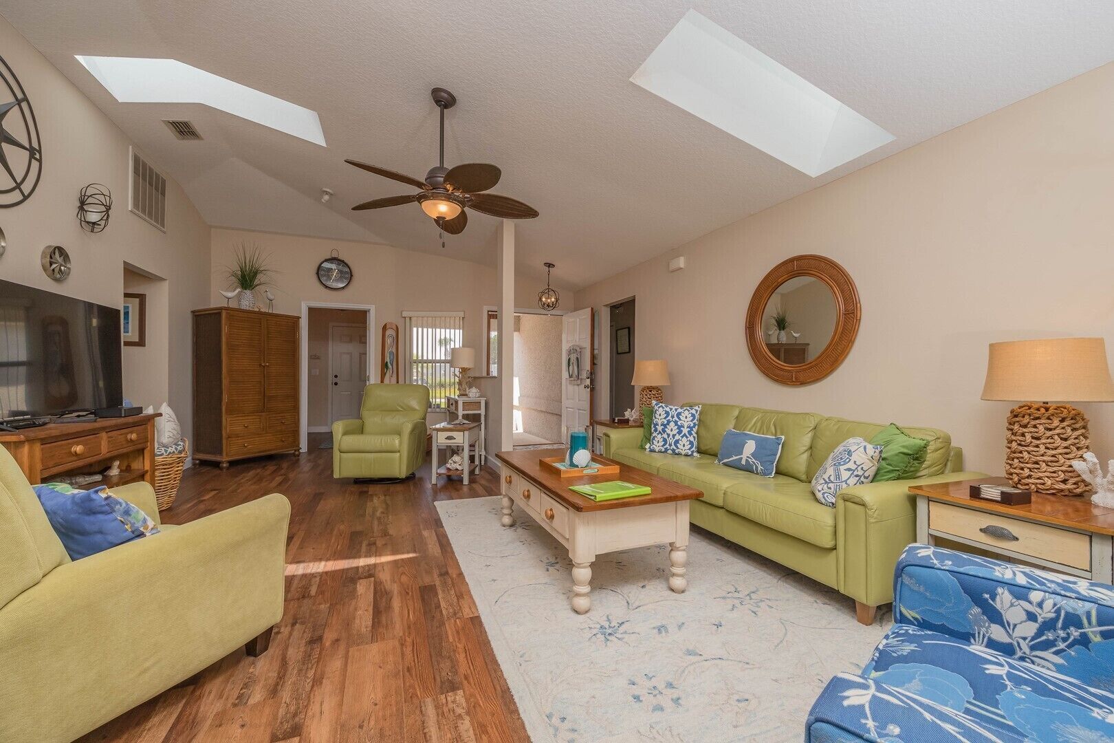 A spacious living room with a wooden floor, a green sofa, a blue patterned sofa, a green armchair, and a wooden coffee table.