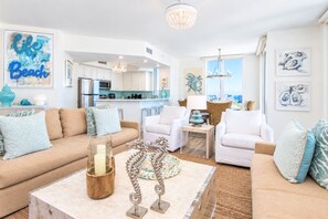 A modern living room with a beige sofa, white armchairs, a coffee table with decorative items, and a kitchen in the background.