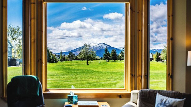 A room with a large window offering a view of a green field, trees, and a mountain. - Golf Course Mountain Home , Views/Hot Tub/Star Gazing/Ski/Hike/Golf/Gather/Relax (Red Lodge)