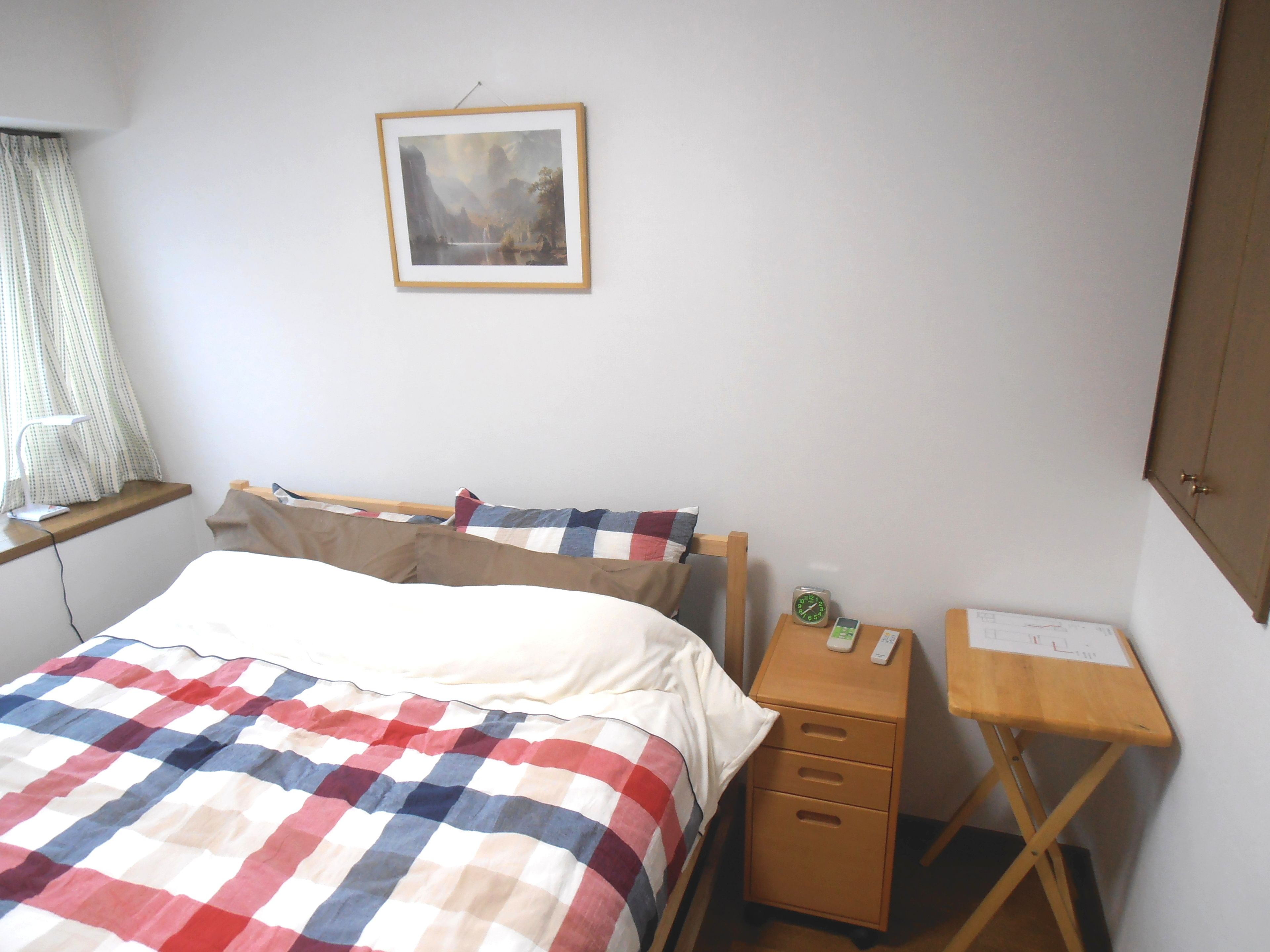 A neatly made bed with a checkered blanket, a bedside table with a clock and a mobile phone, and a framed picture on the wall.