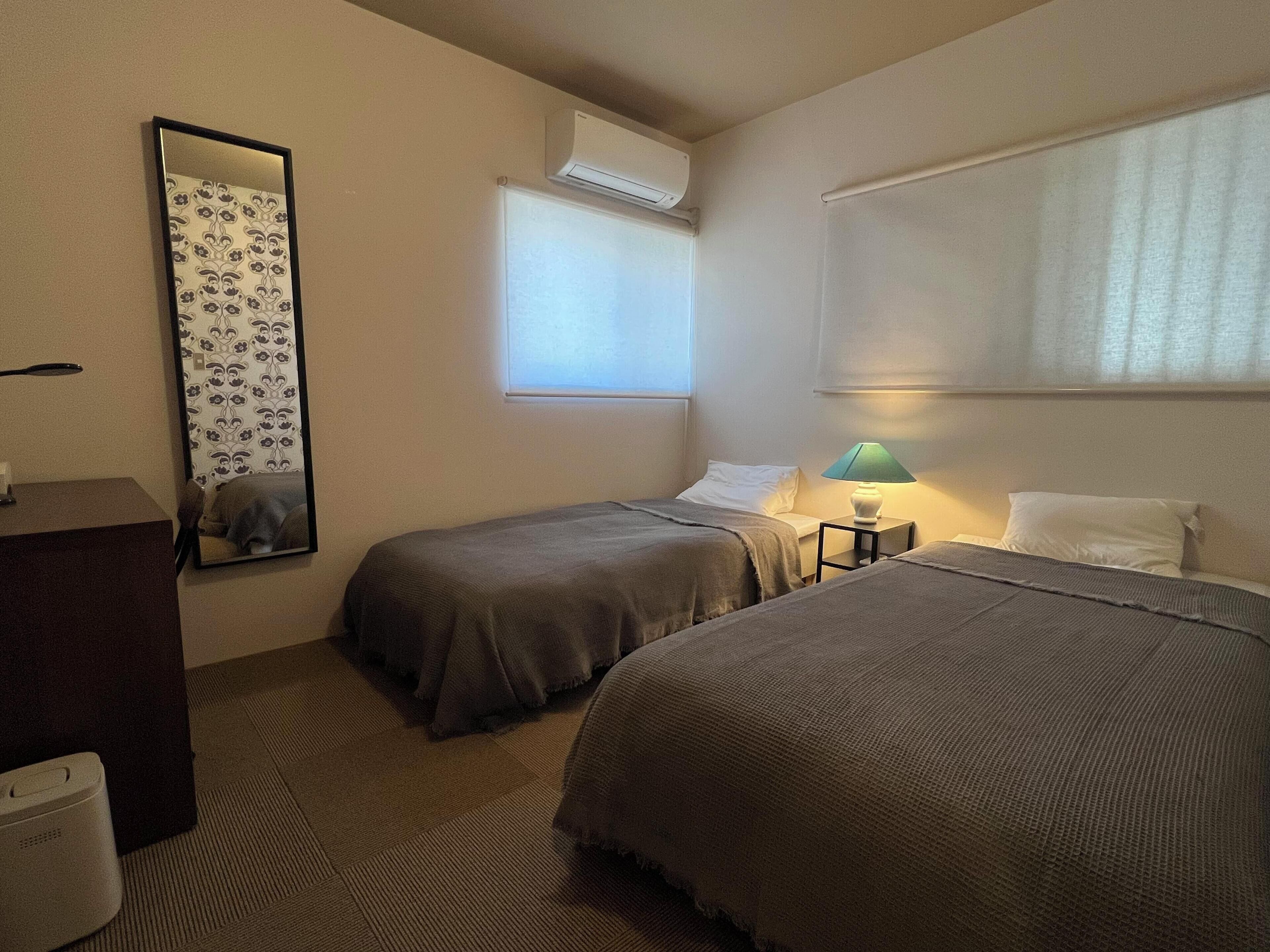 Ein Hotelzimmer mit zwei Einzelbetten, einem Nachttisch mit Lampe, einem Spiegel, einer Klimaanlage und einem Fenster mit Jalousien.
