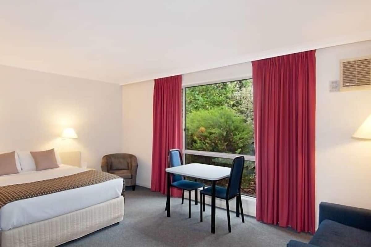 A hotel room with a bed, a desk with two chairs, a window with red curtains, and a view of greenery outside.