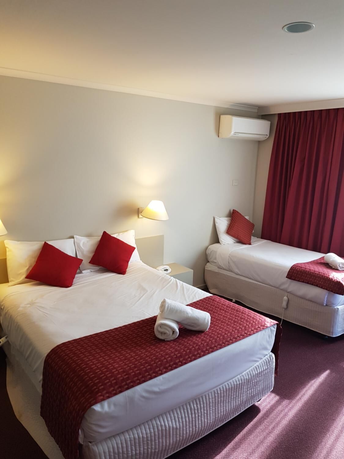 A hotel room with two beds, each with red and white bedding, a red throw blanket, and a white towel on the bed. There is a nightstand with a lamp and a power outlet.
