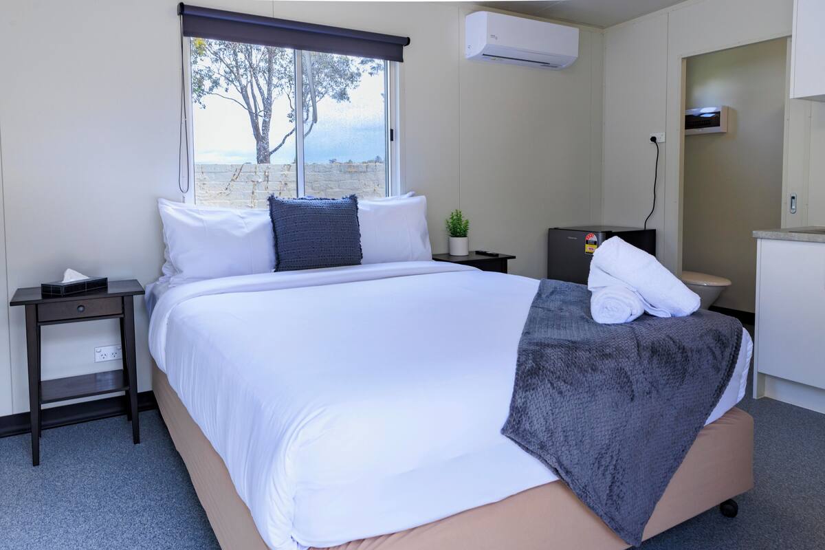 A hotel room with a bed, a small desk, a plant, and a view of the sea.