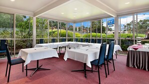 Sala de conferências com mesas e cadeiras, janelas amplas com vista para áreas verdes e edifícios, e teto com iluminação embutida.