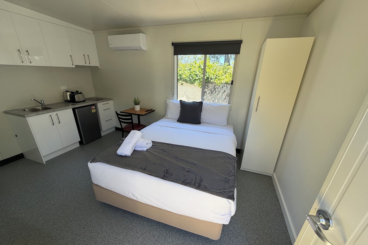 A compact hotel room with a bed, a small desk, a chair, a kitchenette, and a window with a view of greenery.