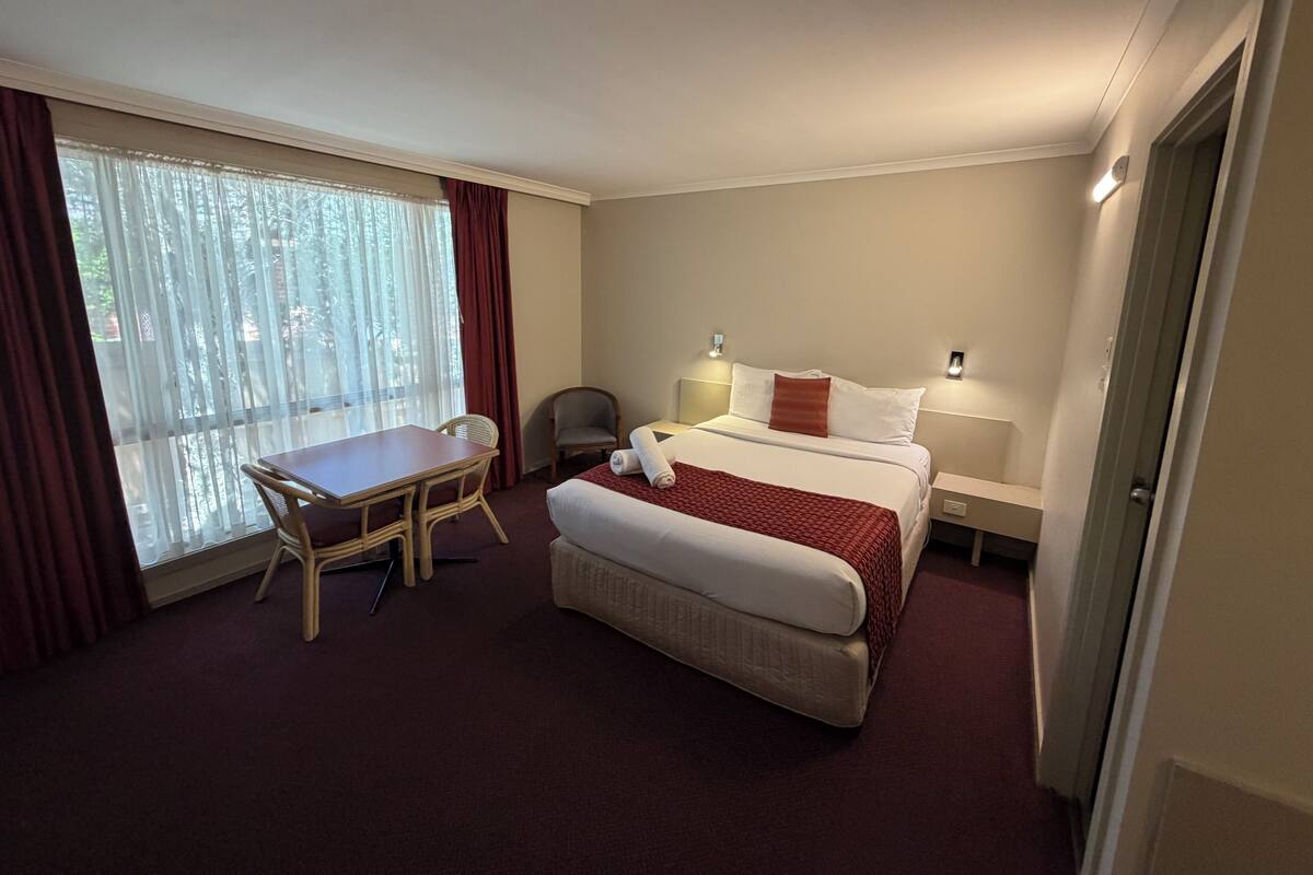 A hotel room with a bed, a desk, and a chair. There is a window with curtains, a nightstand with a lamp, and a door.