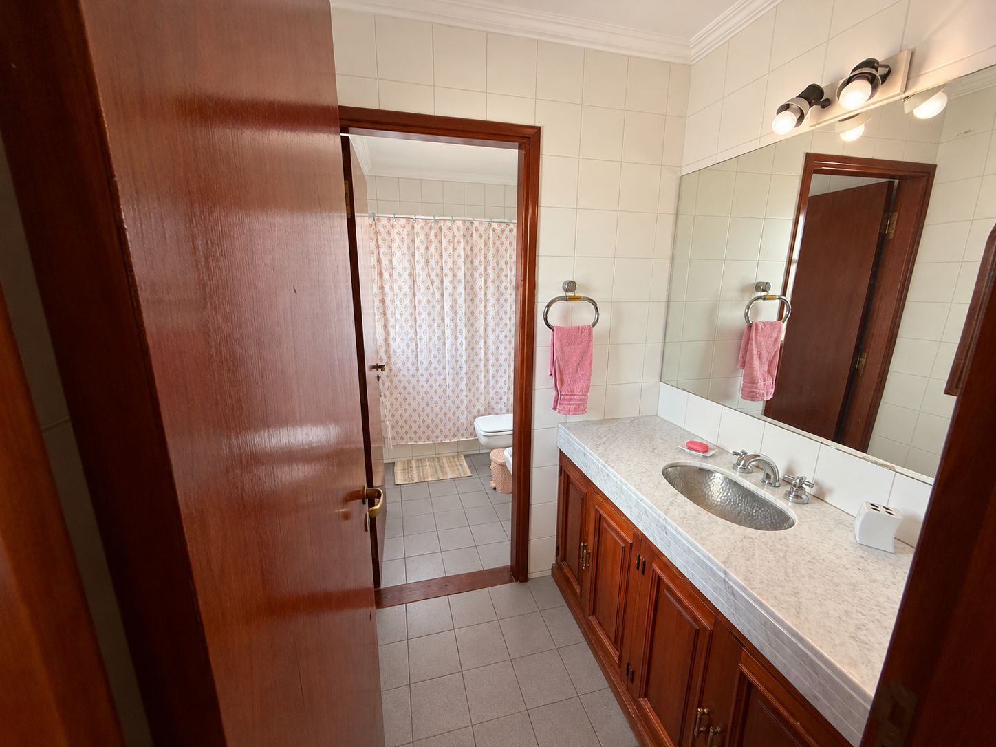 Ein Badezimmer mit Holztür, weißgefliestem Boden, Waschbecken mit Spiegel und Handtuchhalter.