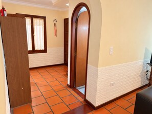 Un passadís amb una porta de fusta, una finestra amb cortines de galeria, una paret blanca amb relleu i un paviment de rajola.
