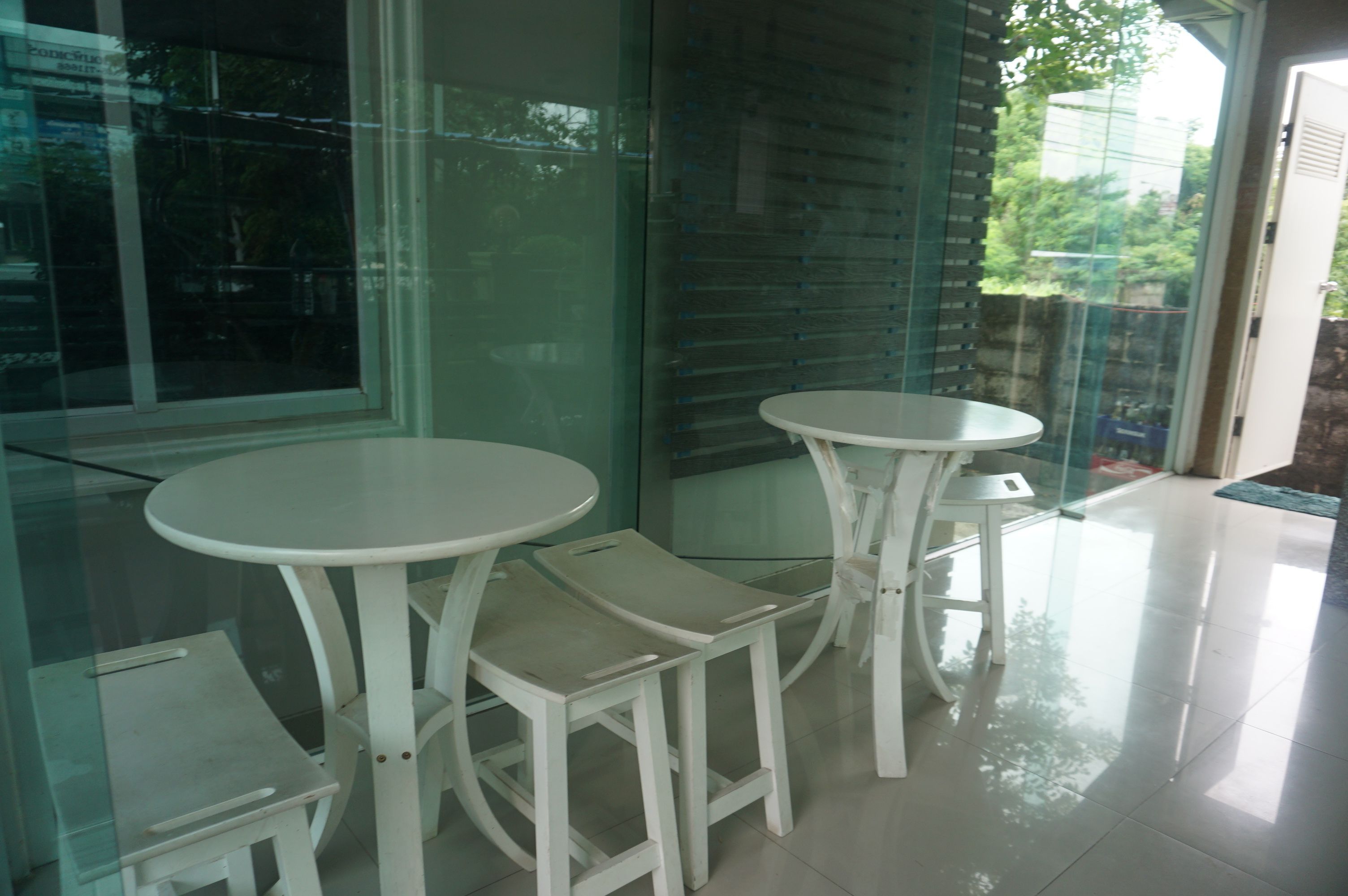 A corner seating area with two white tables and matching chairs, a glass partition, and a view of greenery outside.