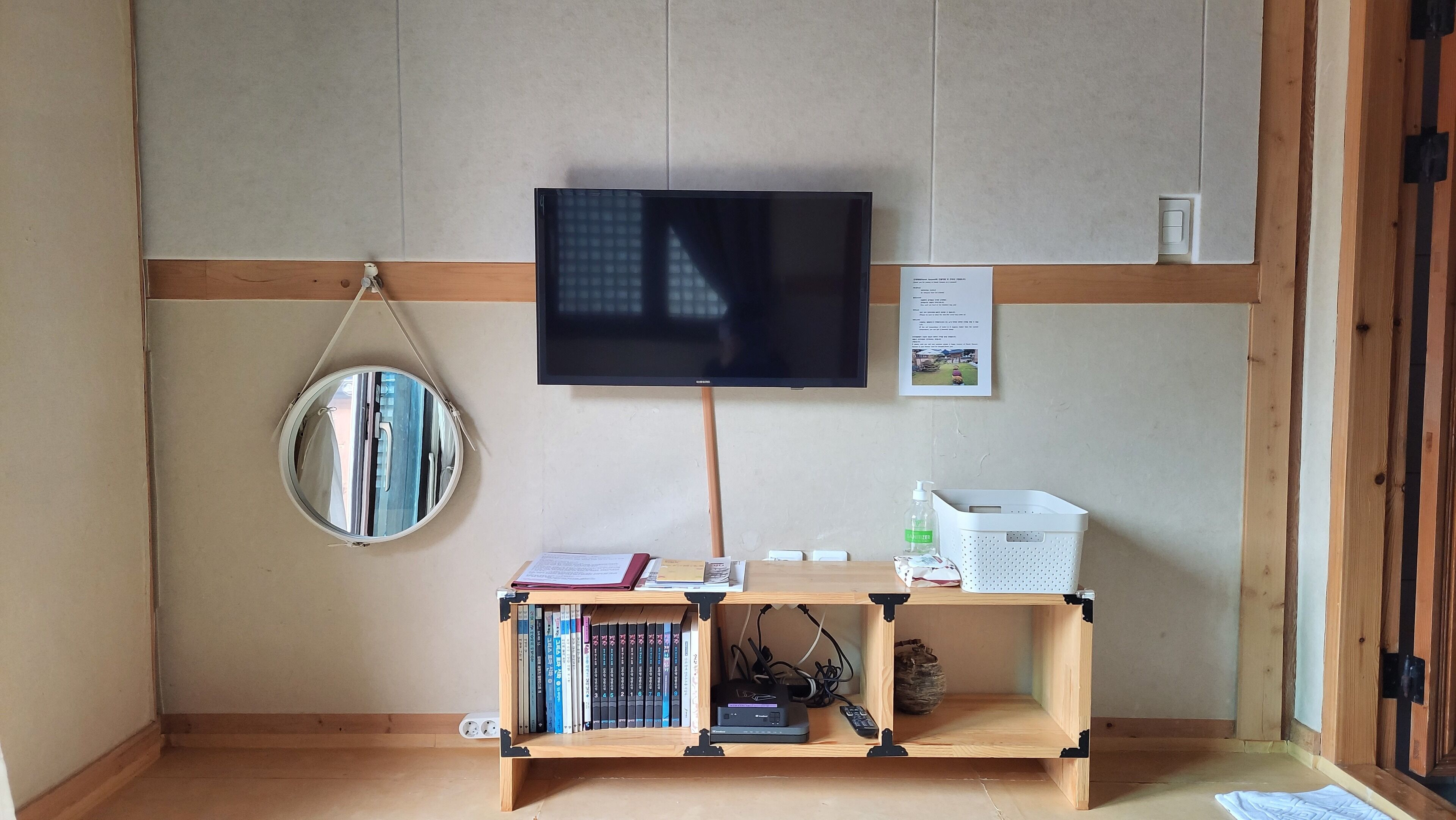 Een kamer met een aan de muur bevestigde televisie, een houten plank met boeken en een kleine speaker, een ronde spiegel en een witte mand.