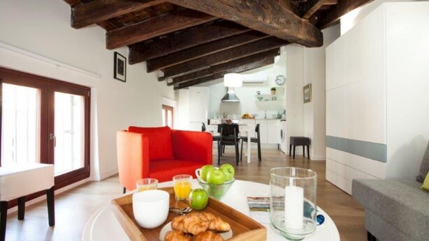 Un plateau de petit-déjeuner avec des croissants, des pommes, du jus de fruits et du lait, disposé dans une pièce dont les poutres en bois sont apparentes, avec un canapé rouge et une vue sur la cuisine.