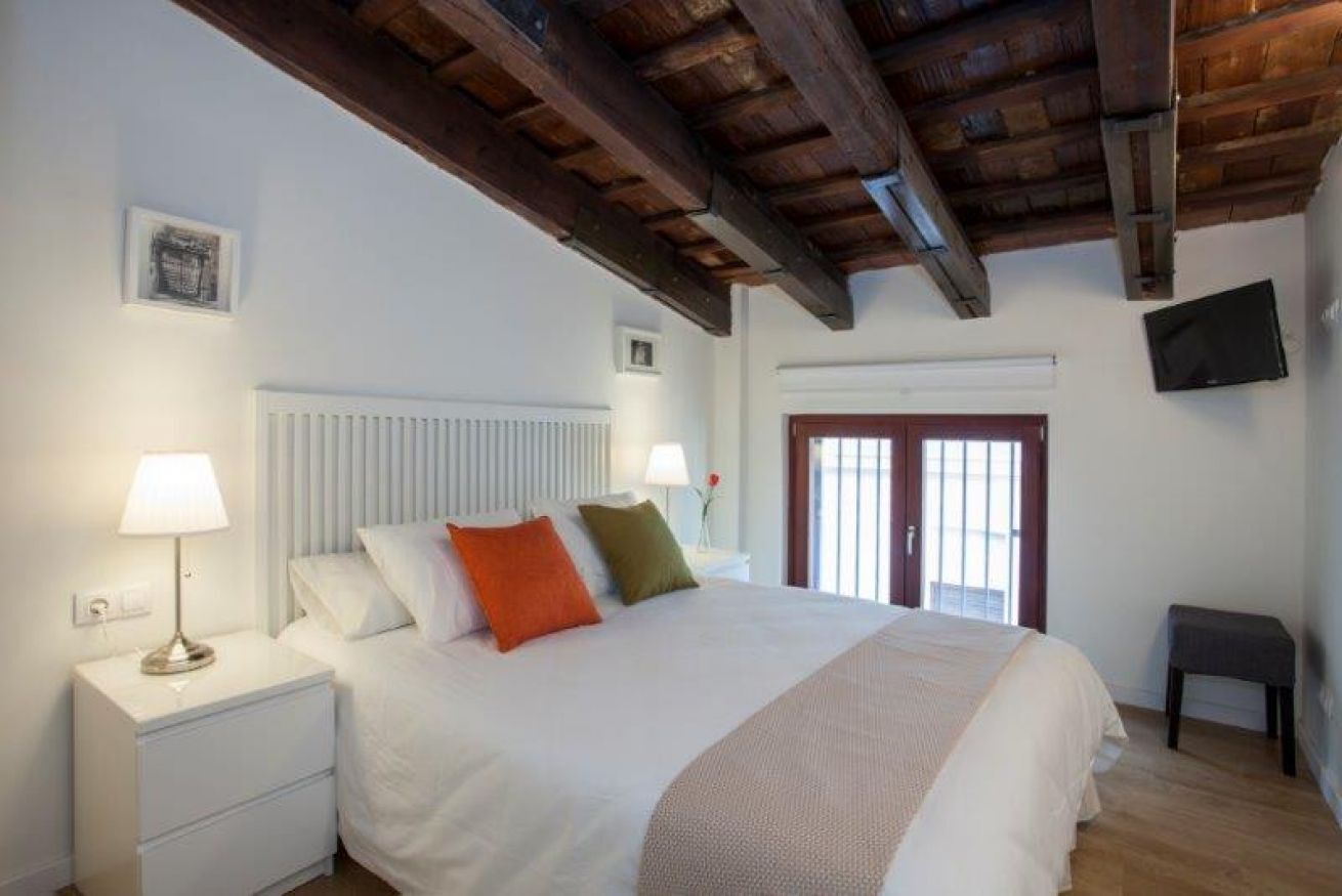 Une chambre avec un grand lit, des tables de chevet blanches, un plafond en bois, un téléviseur fixé au mur et une fenêtre avec des stores.