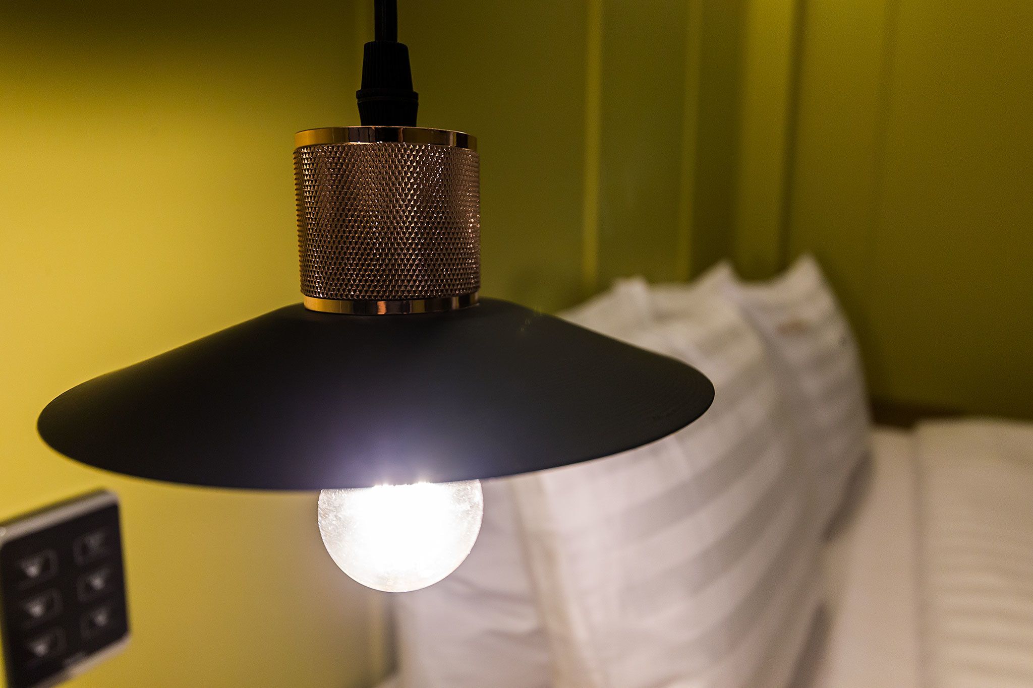 A close-up of a pendant light with a textured lampshade and a visible bulb, hanging in a room with a green wall and white bedding.