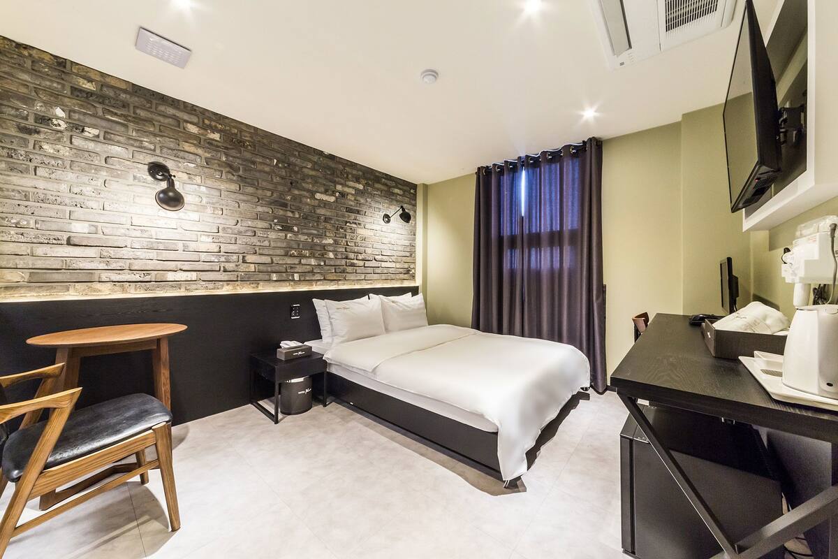 A modern hotel room with a brick-patterned wall, a bed with white linens, a wooden chair, a desk with a coffee maker, and a flat-screen TV mounted on the wall.