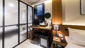 A compact hotel room with a desk, a small refrigerator, a bed with pillows labeled "Hotel Plum," and a window with blinds. - Hotel Plum (Siheung)