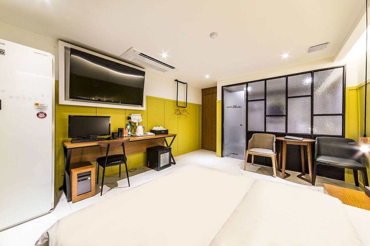 A compact hotel room with a bed, a desk with a computer, a chair, a TV, and a window with a view.