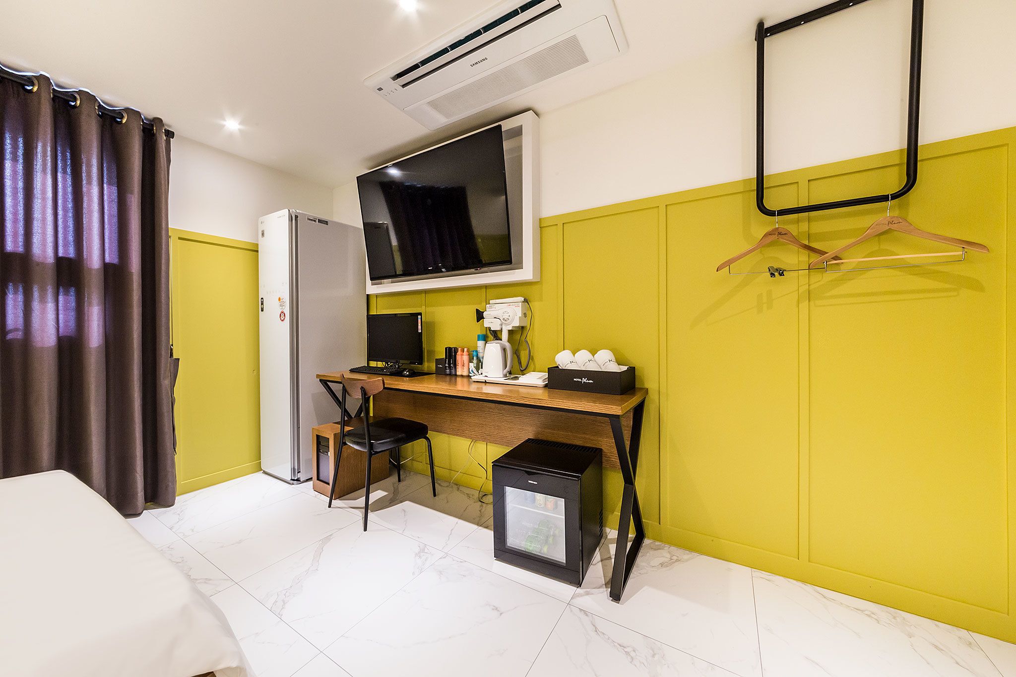 Habitación de hotel pequeña con cama, escritorio con computadora, refrigerador, televisor y perchero.
