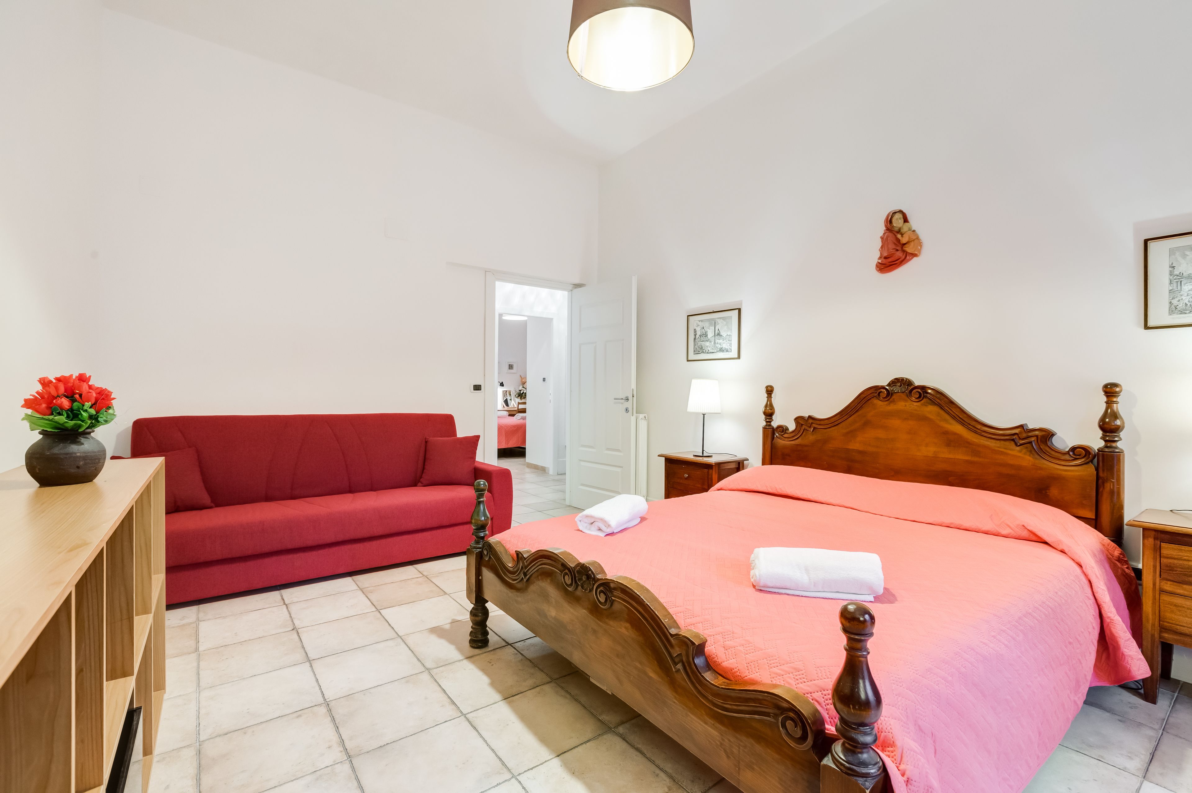 Una habitación de hotel con una cama de madera, un sofá rojo, una mesita de noche de madera y un jarrón con flores.