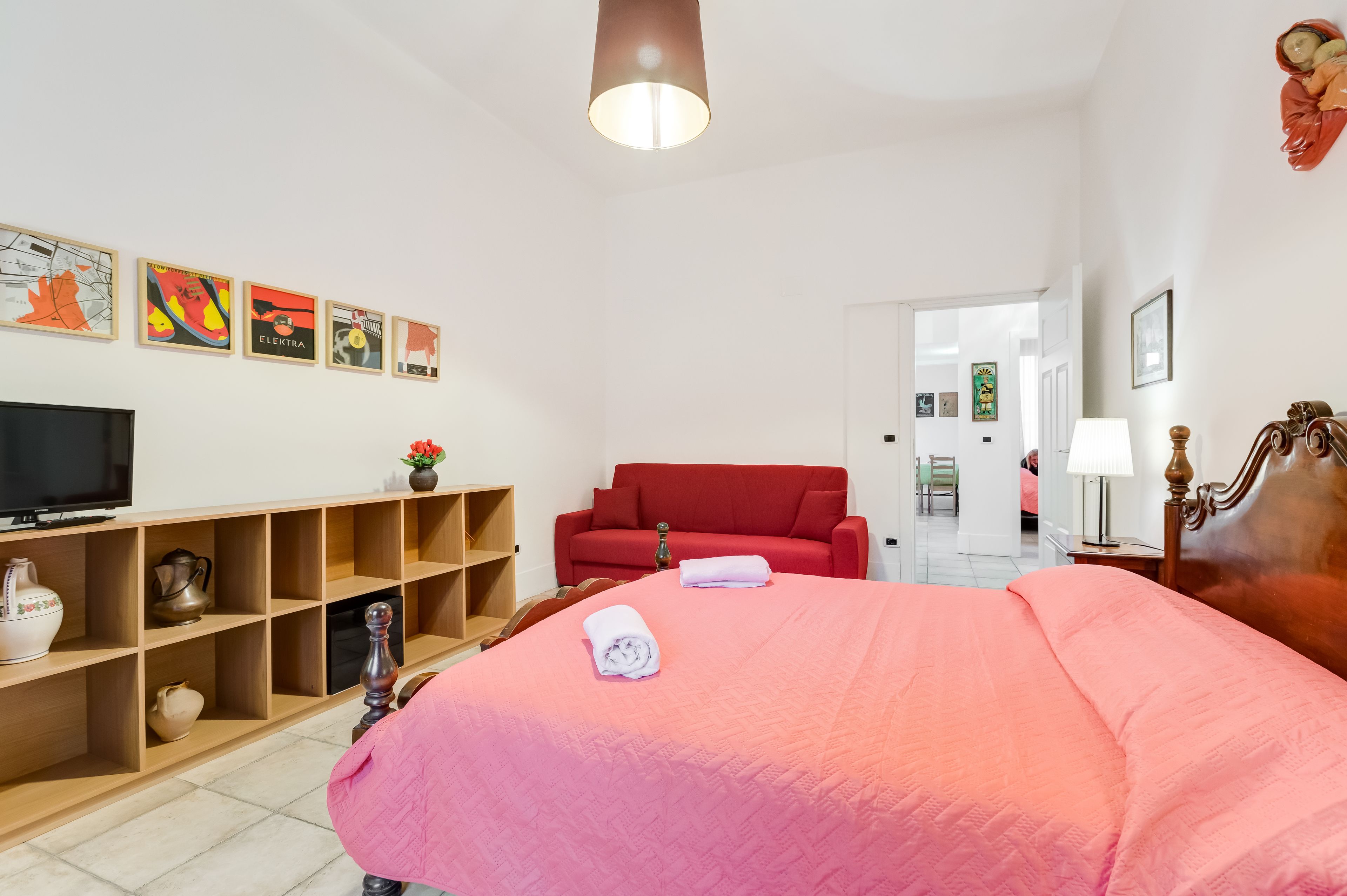 Una habitación de hotel con una cama, un sofá rojo, un televisor y una estantería con objetos decorativos.
