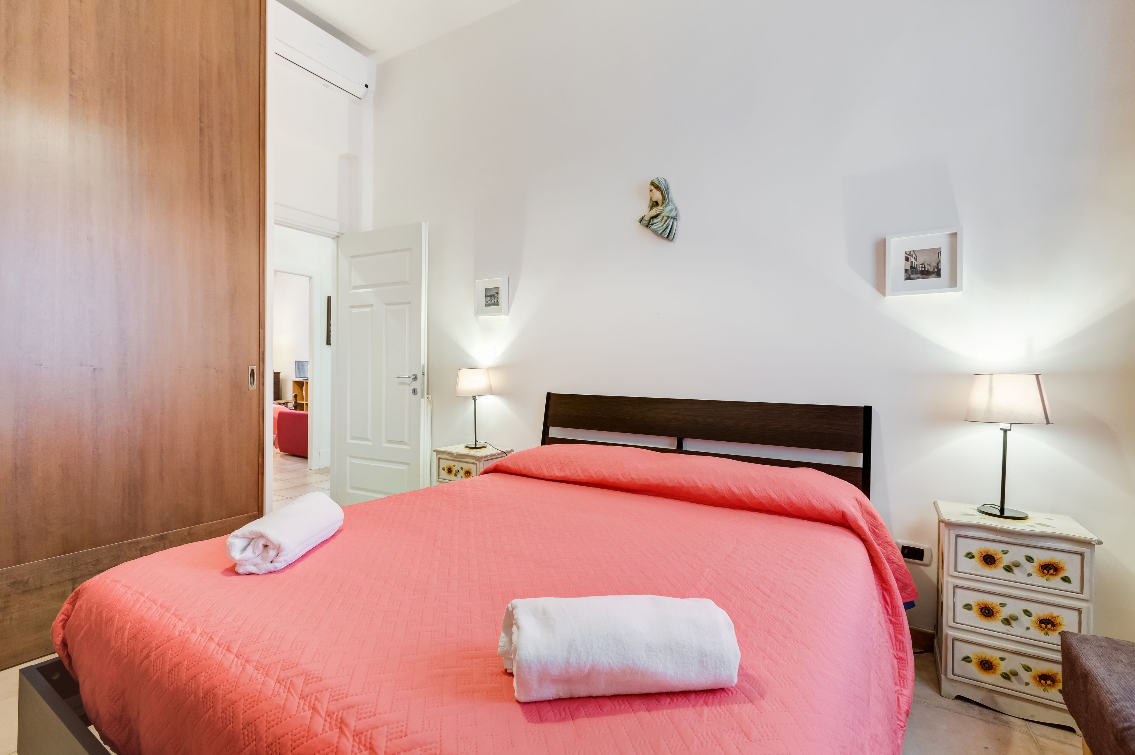 Ein ordentlich bezogenes Bett mit rosa Bettwäsche und weißen Handtüchern, ein Holzschrank, ein Nachttisch mit Lampe und ein an der Wand hängendes Bild.