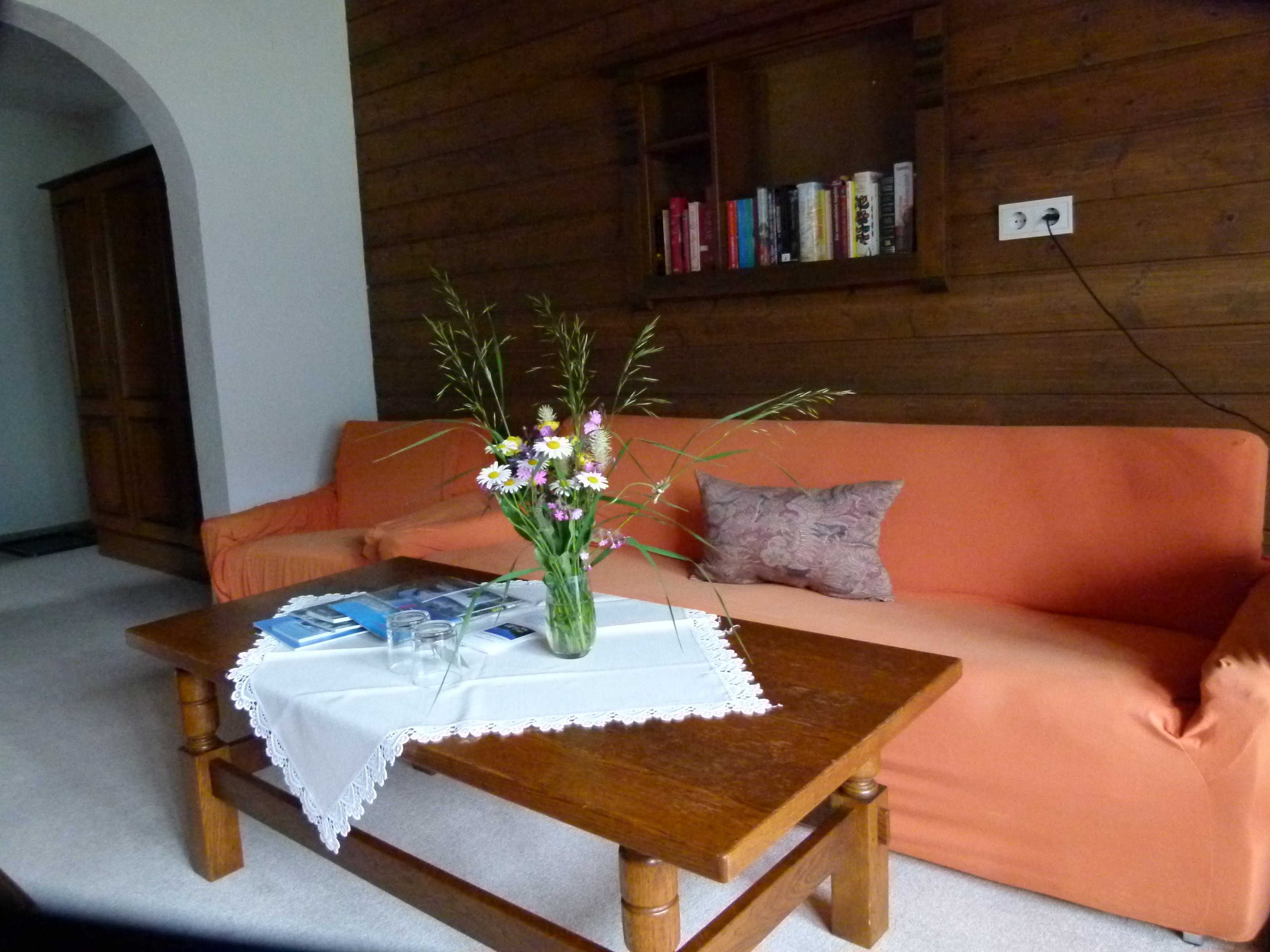 A wooden interior with a wooden table, a white tablecloth, a vase with flowers, a sofa, and a bookshelf.