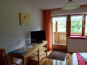 Une pièce avec un bureau en bois, une télévision, une fenêtre avec des rideaux et une vue sur la verdure à l’extérieur.