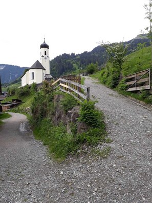 A gravel path leading to a white church with a bell tower, surrounded by greenery and hills. - Double room new max 2P (type A) (copy) Z-NR. 5 - Greber Christine - (Schoppernau)