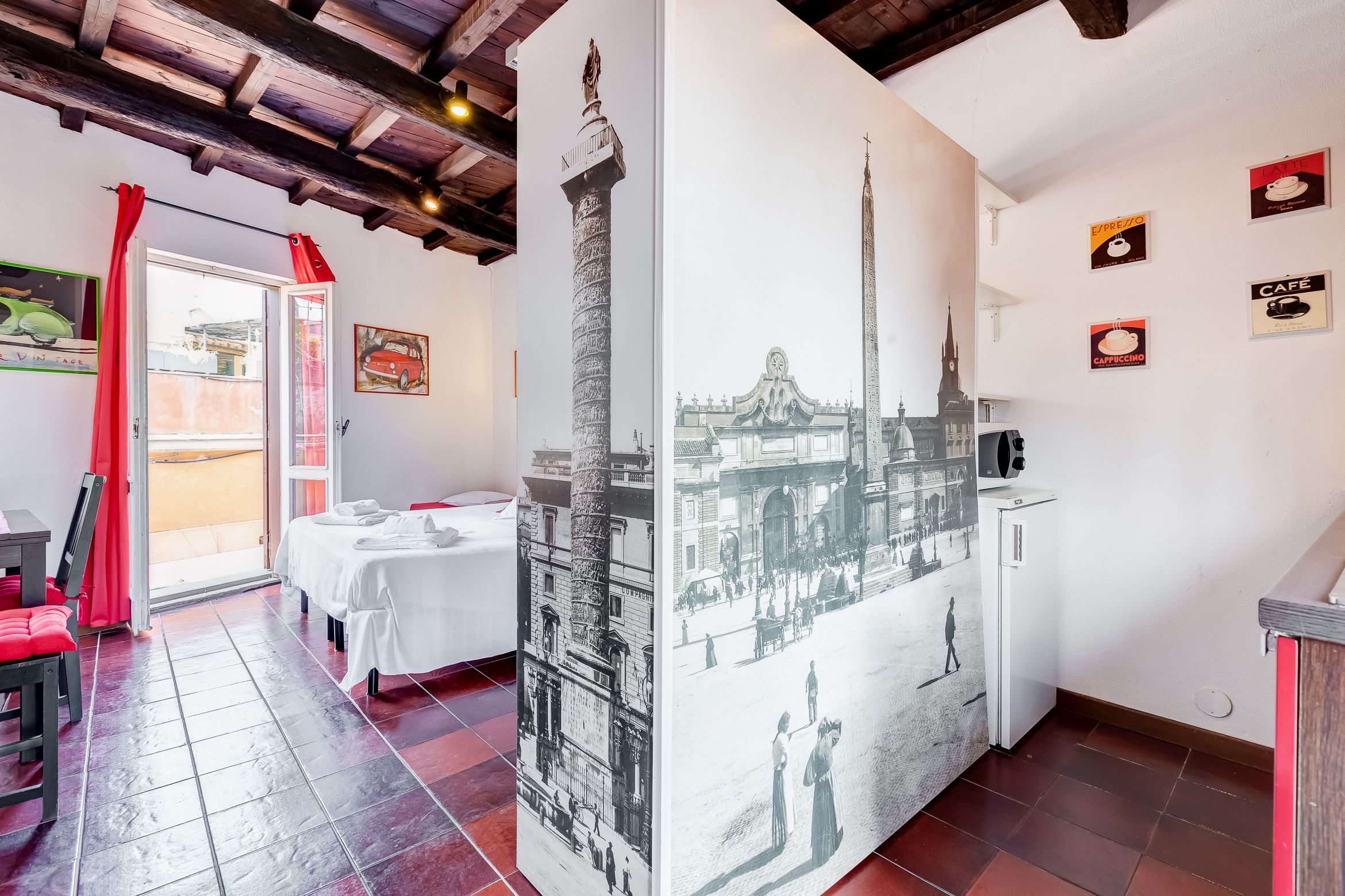 Habitación de hotel con cama, mesa de comedor y kitchenette. Hay carteles en las paredes y una ventana con cortinas rojas.