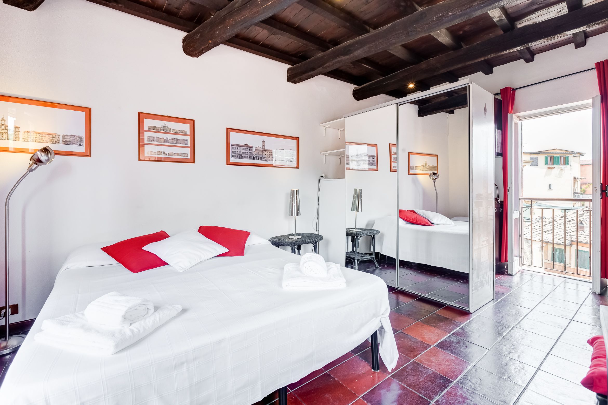 A hotel room with a bed, a mirror, a red chair, and a balcony with a view of buildings.