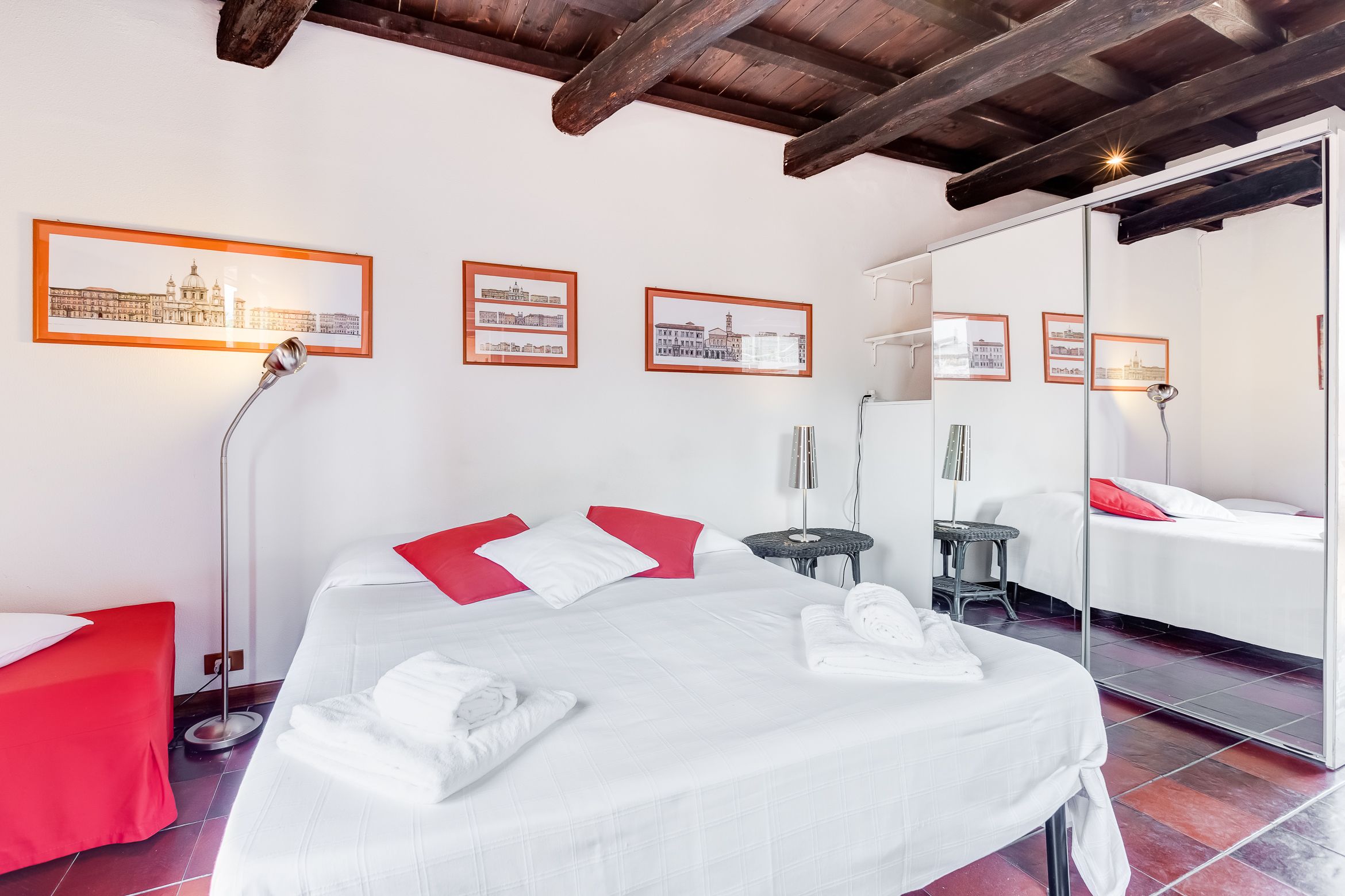 A hotel room with a large bed, red and white pillows, a red chair, a small table, a floor lamp, and framed pictures on the wall.