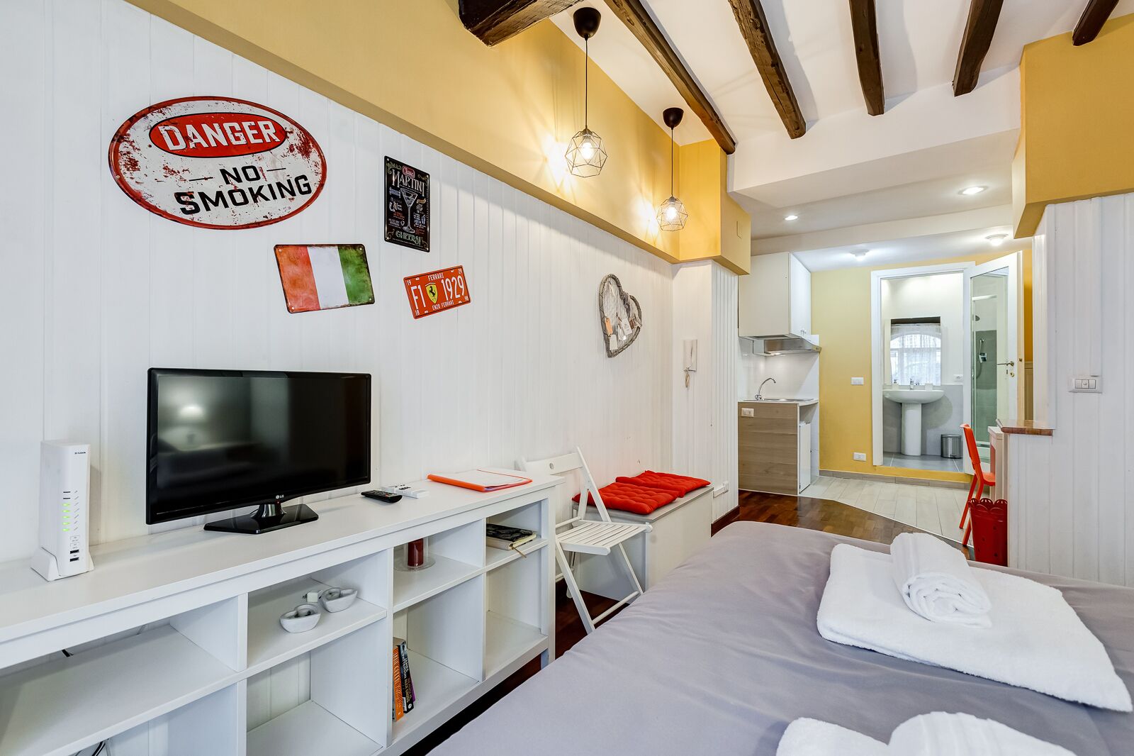 A modern bedroom with a TV, storage shelves, a bed with a red pillow, and a bathroom visible through an open door.