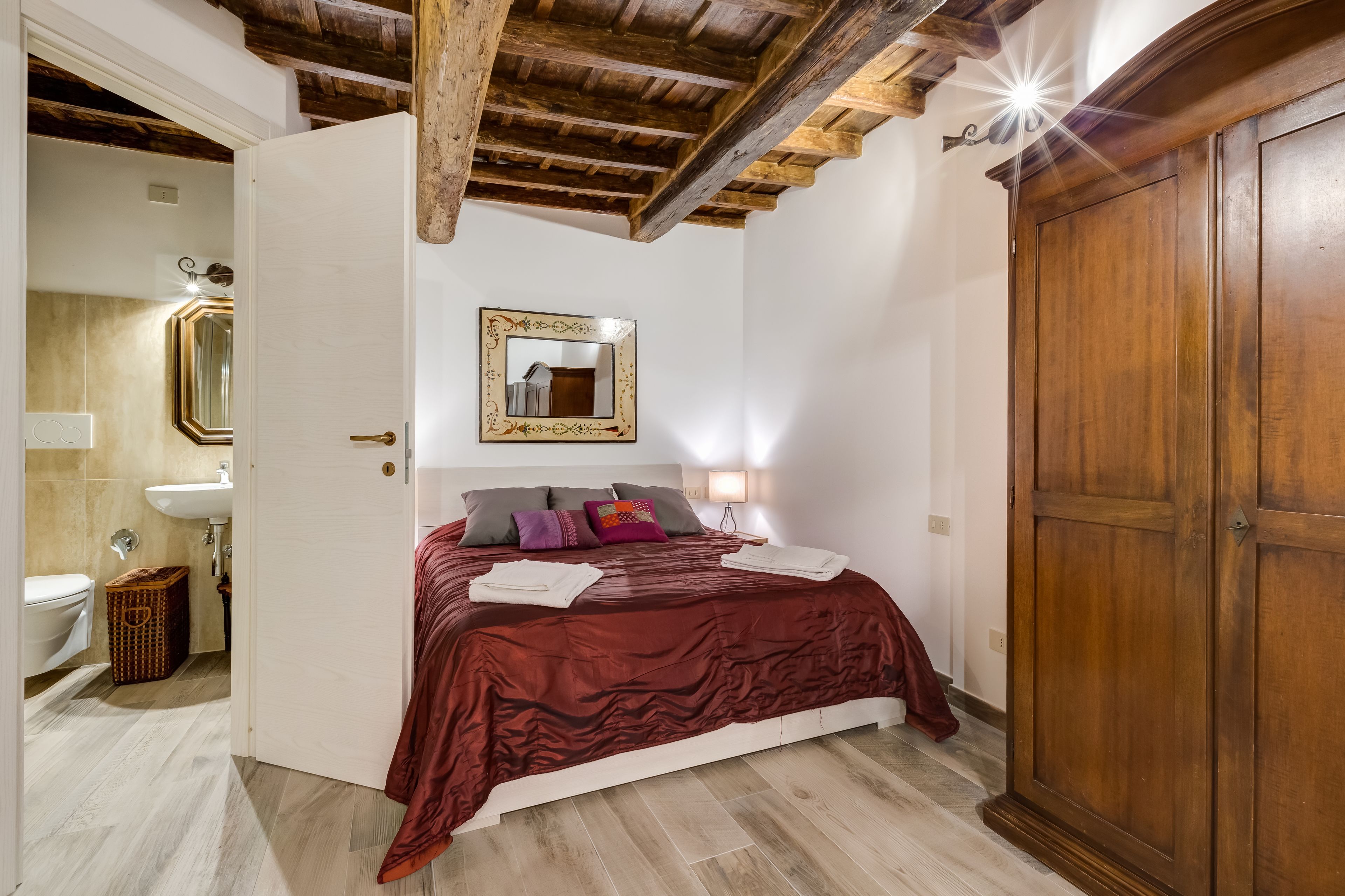 Une chambre avec un plafond en bois, un lit recouvert d’une couverture rouge, une table de chevet avec une lampe et une armoire.