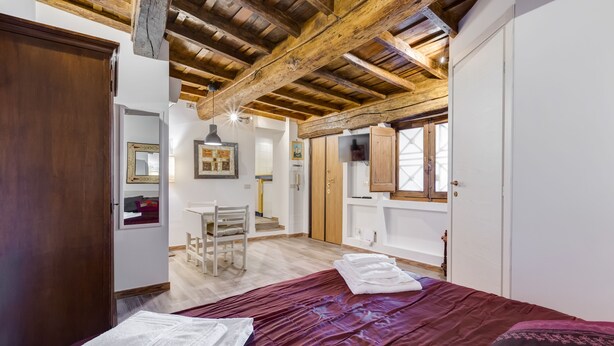 A bedroom with a wooden ceiling, a bed with a red bedspread, a white chair, and a small desk with a chair. - Pellegrino Campo de' Fiori Suite (Rome)