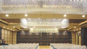A banquet hall with rows of white chairs with blue sashes, a chandelier, and a patterned carpet. - Hotel Grand Maple (Jaipur)