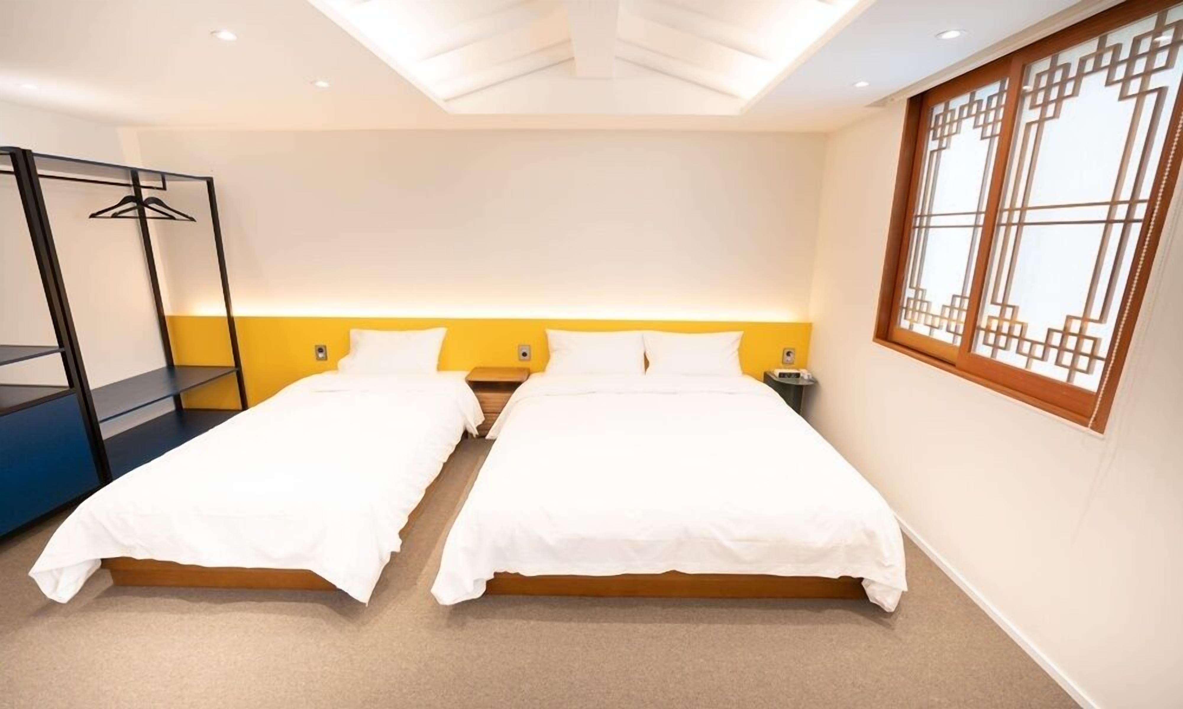 Quarto de hotel com duas camas, um guarda-roupa, uma mesa de cabeceira e uma janela com padrão geométrico.