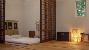 Ein traditioneller japanischer Raum mit Tatami-Boden, niedrigem Bett, Shoji-Schirm, kleinem Tisch mit Lampe und Fenster mit Blick auf Grünflächen.