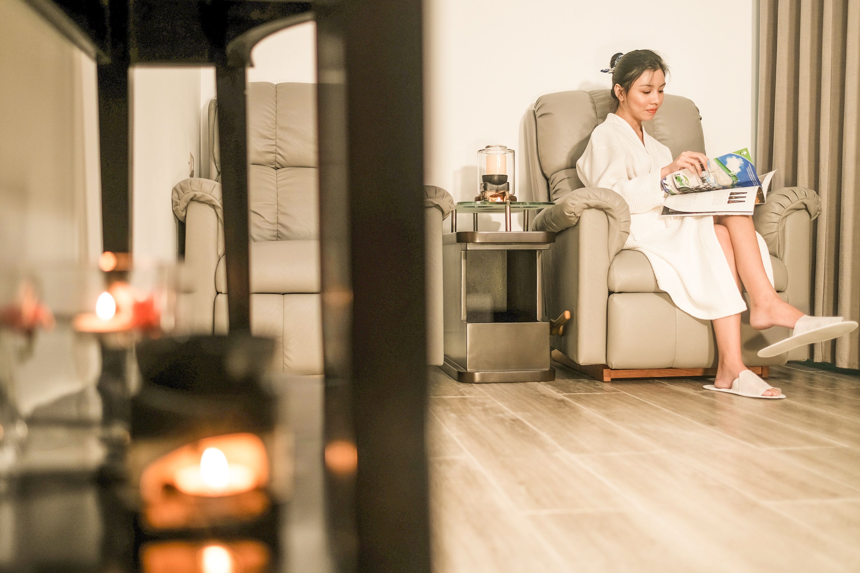 Uma pessoa vestida com roupeiro sentada numa cadeira de massagem num quarto com uma mesa de massagem, uma mesinha com uma vela e um recipiente de vidro.