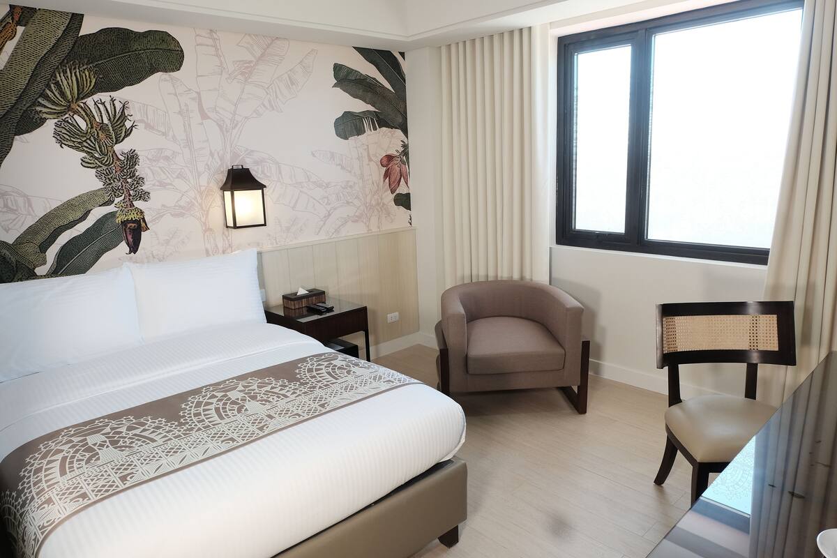 Una habitación de hotel con una cama grande, una silla, un escritorio, una lámpara y un ventanal con cortinas.