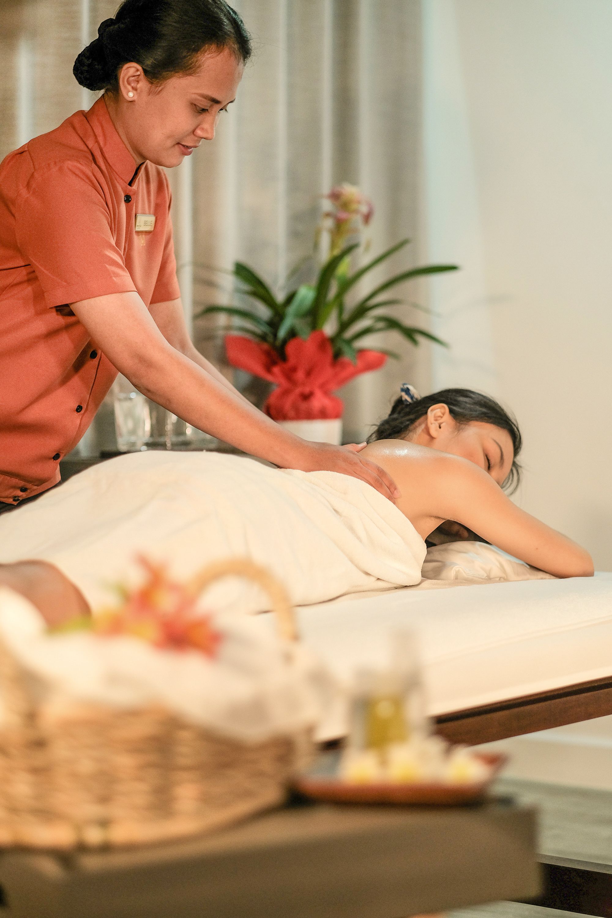 A massage therapy session in a spa setting with a client lying on a massage table, a therapist attending to the client, and a decorative vase with flowers in the background.