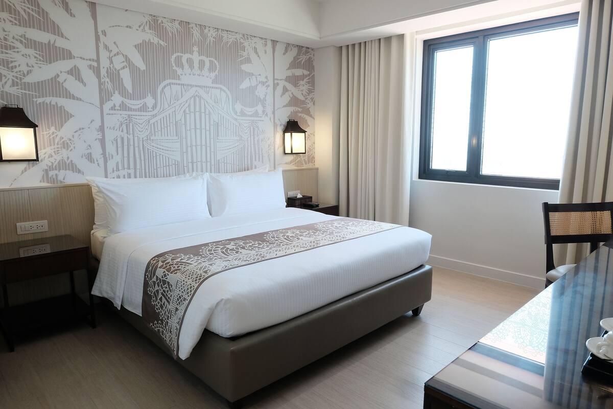 Habitación de hotel con una cama grande, dos mesitas de noche con lámparas, una ventana con cortinas y vistas al exterior.