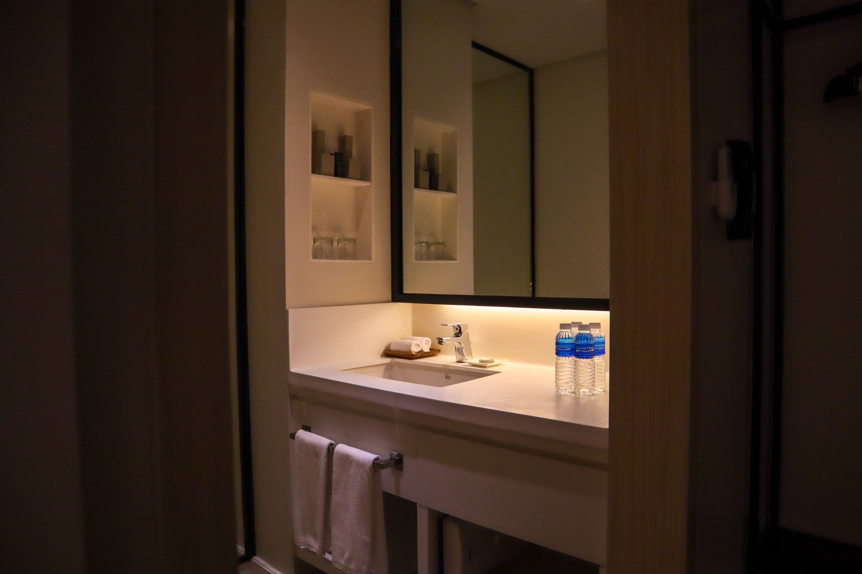 Un baño con un lavabo blanco, un espejo y repisas con artículos de tocador.