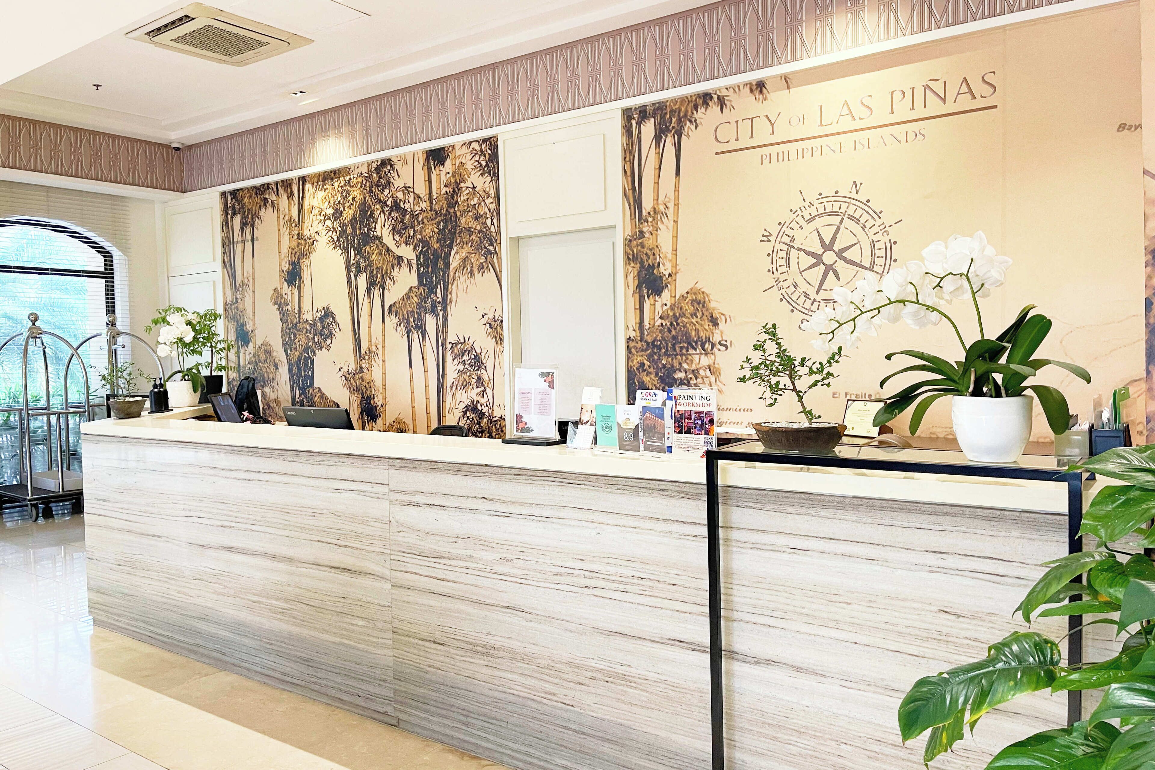 A reception desk with a wooden finish, a large wall mural depicting palm trees and a compass, and several potted plants.