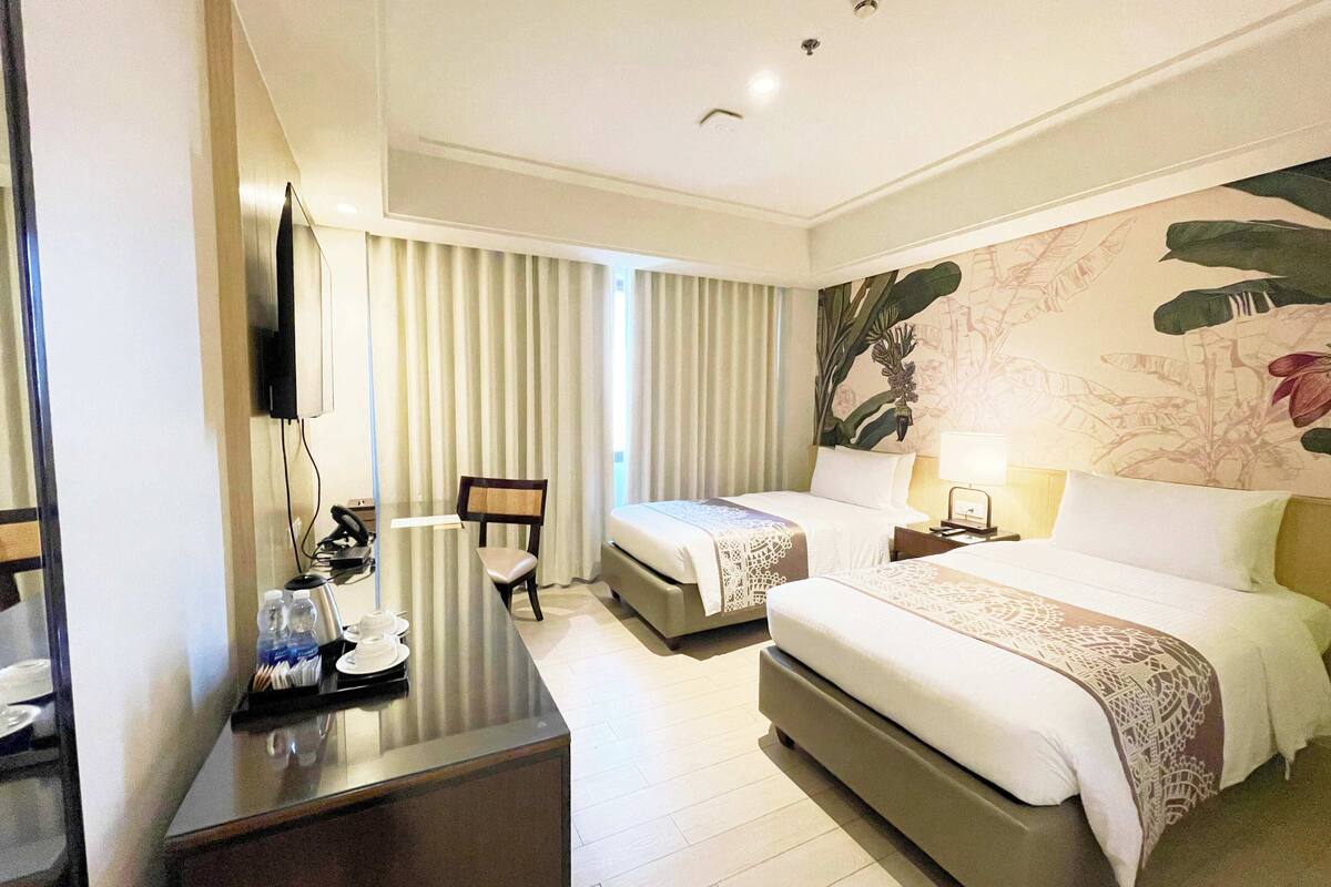 Habitación de hotel con dos camas, un escritorio con silla, televisión y un ventanal con cortinas.