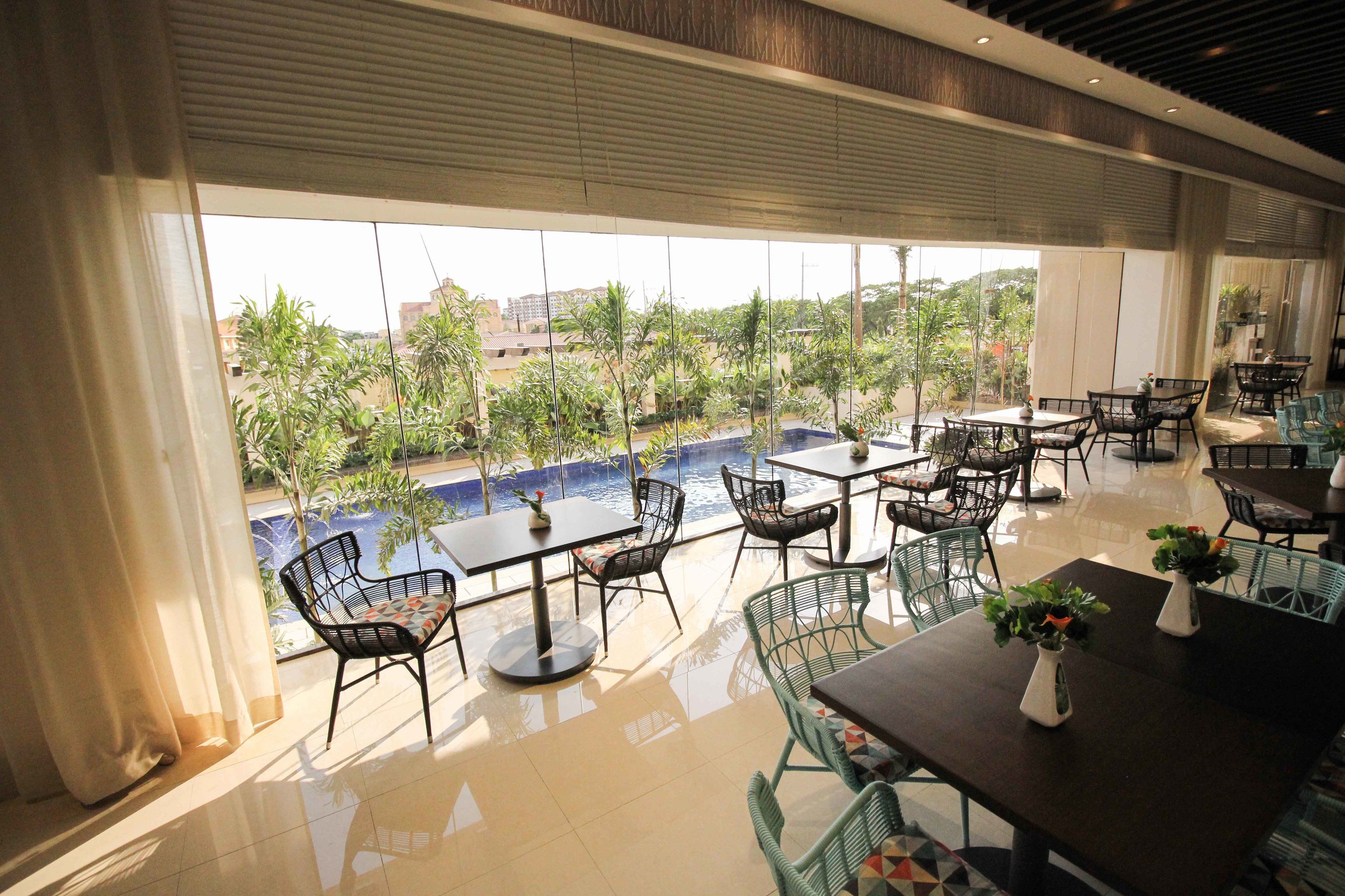 A spacious outdoor dining area with tables and chairs, large windows offering views of a swimming pool and greenery.
