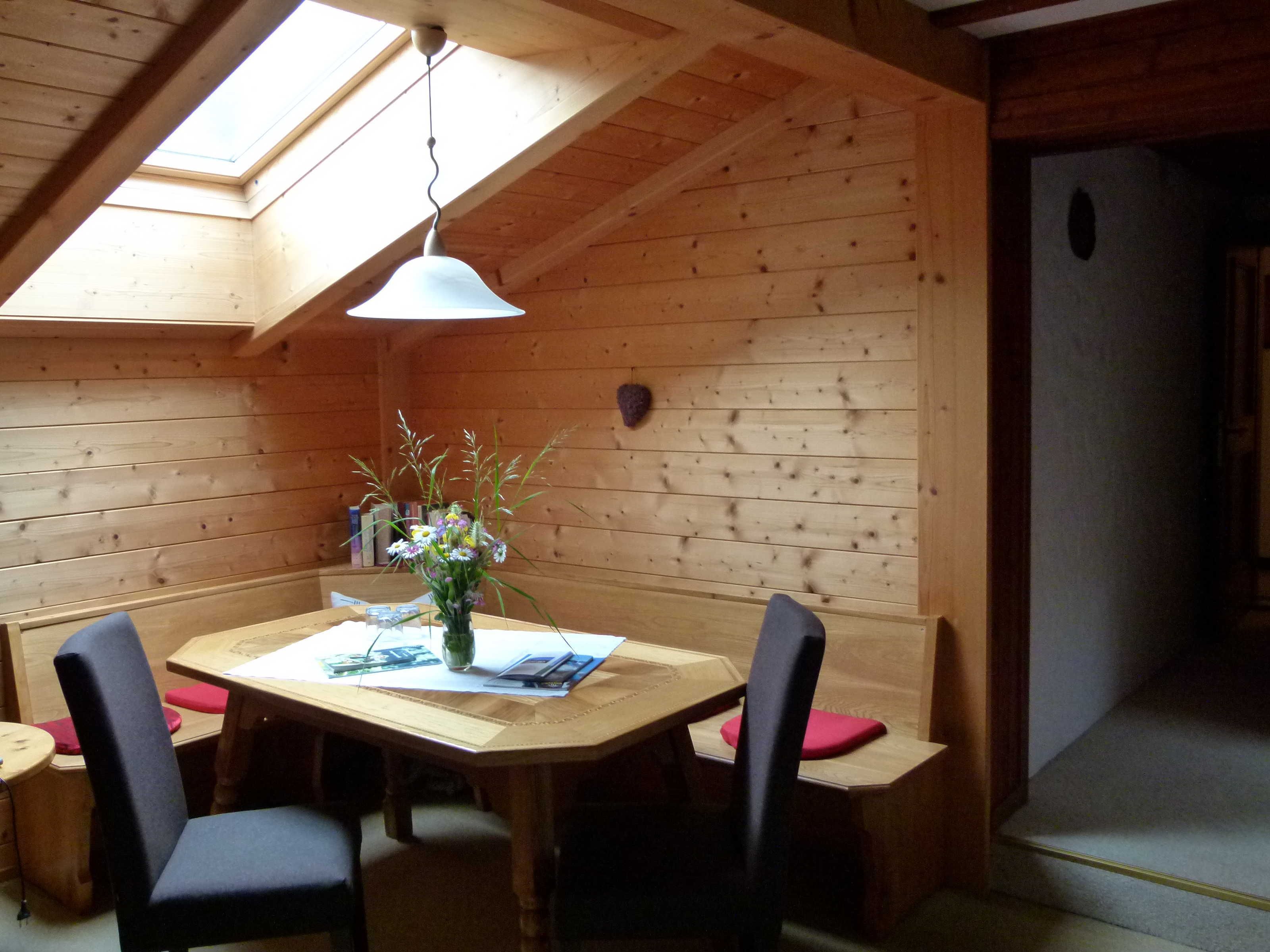 Un intérieur en bois avec un plafond haut, une lucarne, une table avec des chaises et un vase de fleurs.