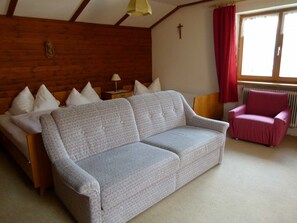 A room with a grey sofa, a wooden headboard, a wooden floor, a red chair, a window with curtains, and a cross on the wall. - Triple Room (Type A) - Greber Christine - Frühstückspension Fernblick (Schoppernau)