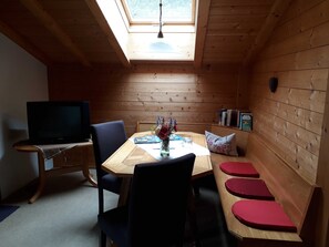 A cozy room with a wooden interior, a TV on a stand, a dining table with chairs, and a bench with red cushions. - Triple Room (Type A) - Greber Christine - Frühstückspension Fernblick (Schoppernau)