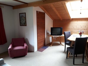 A cozy room with a TV on a wooden stand, a dining table with chairs, a red armchair, and a wooden ceiling. - Triple Room (Type A) - Greber Christine - Frühstückspension Fernblick (Schoppernau)