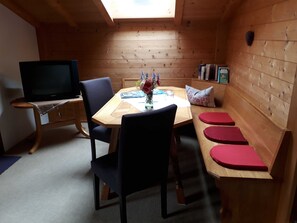 A cozy room with a wooden table, chairs, a TV, and a small bench with red cushions. - Triple Room (Type A) - Greber Christine - Frühstückspension Fernblick (Schoppernau)